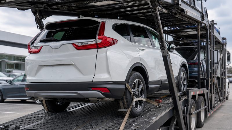 A white Honda CR-V secured with wheel straps on the lower deck of an open auto transport carrier in a dealership lot