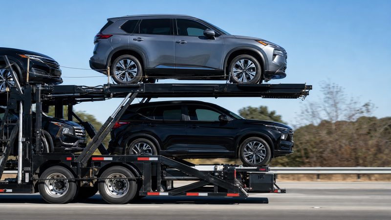 A grey Nissan Rogue on the top deck of a loaded open auto transport carrier on the interstate