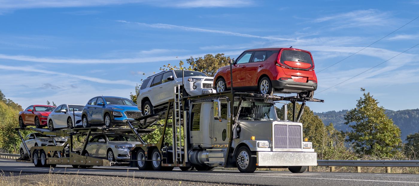 An open car carrier photographed side on: a stinger tractor with a head rack over the cab pulling a two-deck open trailer loaded with cars